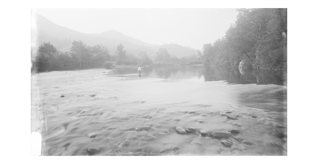 Black and white image of a man standing in a river with mountains in the background. 