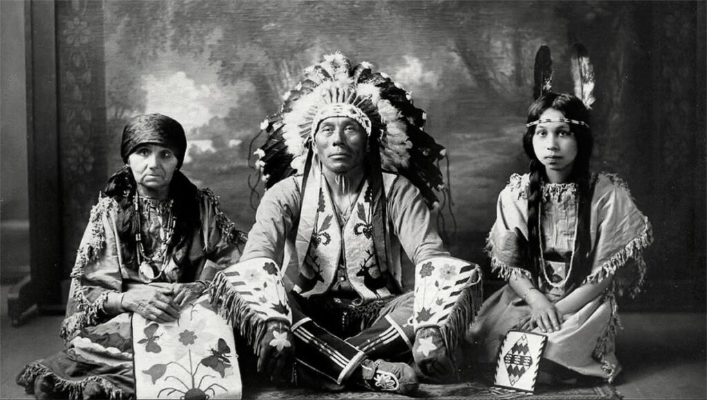 Chief Shelton, his wife, and daughter all wearing traditional regalia in a black and white photograph. Chief Shelton is wearing a large war bonnet and his daughter and wife are displaying handicrafts. 
