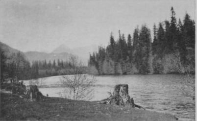 Black and white photograph (artist unknown) of a river bordered by pine trees and mountains. 