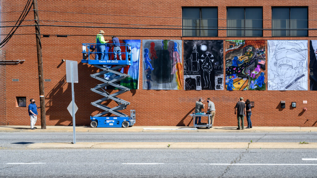 This photograph shows a tall brick wall on the side of a building. To the left of the photo, workers are installing large banners on the wall using a scissor lift, a piece of construction machinery that raises a platform high into the air so that work can be performed off the ground. To the right of the photo, a small group of people are installing plaques onto the wall below the banners. The photograph was taken across the street from the wall.
