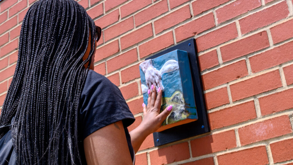 This photograph is a close-up of Industries of the Blind employee Tanessa Brown touching a ceramic relief sculpture as part of an art installation based on her as the subject of conversations with a painting student. The photograph shows Tanessa from the back, with her left hand outstretched to touch the sculpture. She has long box braids extending down her back. The finger nails on her outstretched hand are bright pink. The ceramic sculpture depicts a hand reaching down from the sky, which is blue with white clouds. 