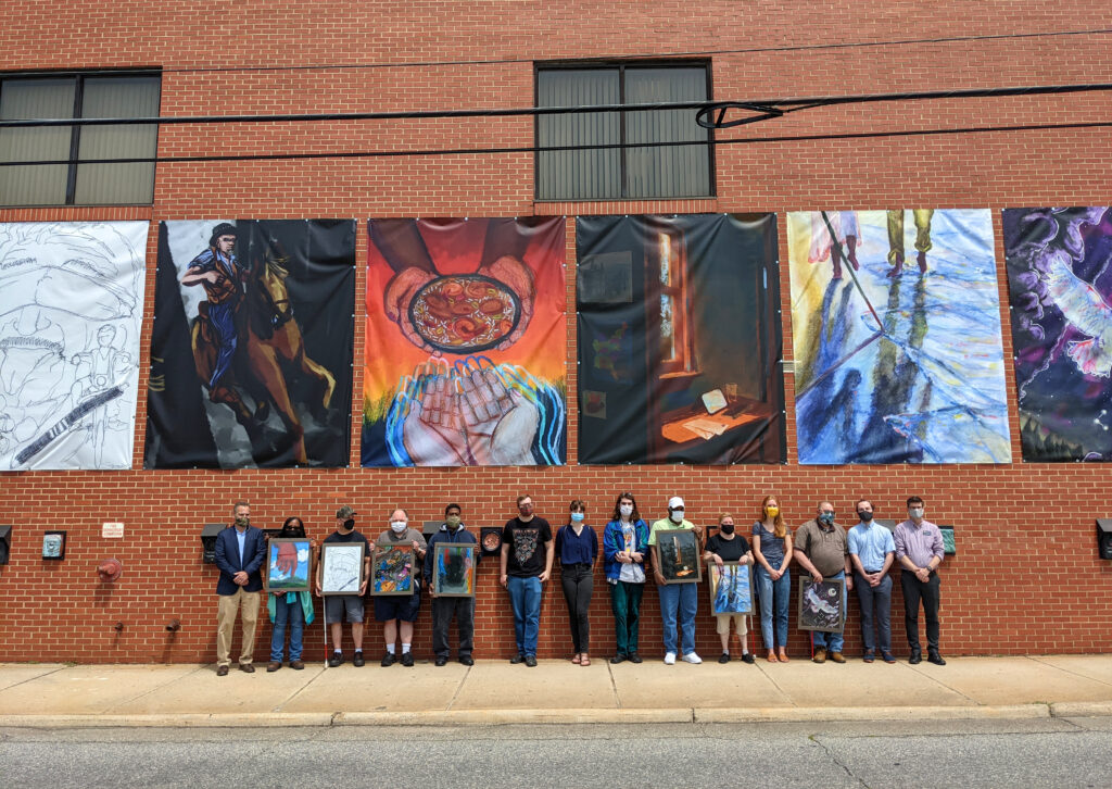 This photograph shows a view of six large banners installed on a tall, red brick wall. The banners show art works with vibrant colors and a range of compositions, from dynamic figures to quiet interior settings. Below the banners stand a group of fourteen people, side-by-side, of different heights, ages, and races.